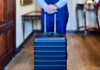Schwarzer Koffer auf Rollen, am Ausziehgriff gehalten von einem Butler in blauer Hose und Weste.