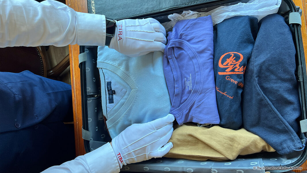 Ein geöffnet liegender Rollkoffer mit gefalteten T-Shirts und Pullovern. Die weißen Handschuhe eines Butlers packen die Wäsche über den Schuhen in den Koffer, die direkt über den Rollan gelagert sind.